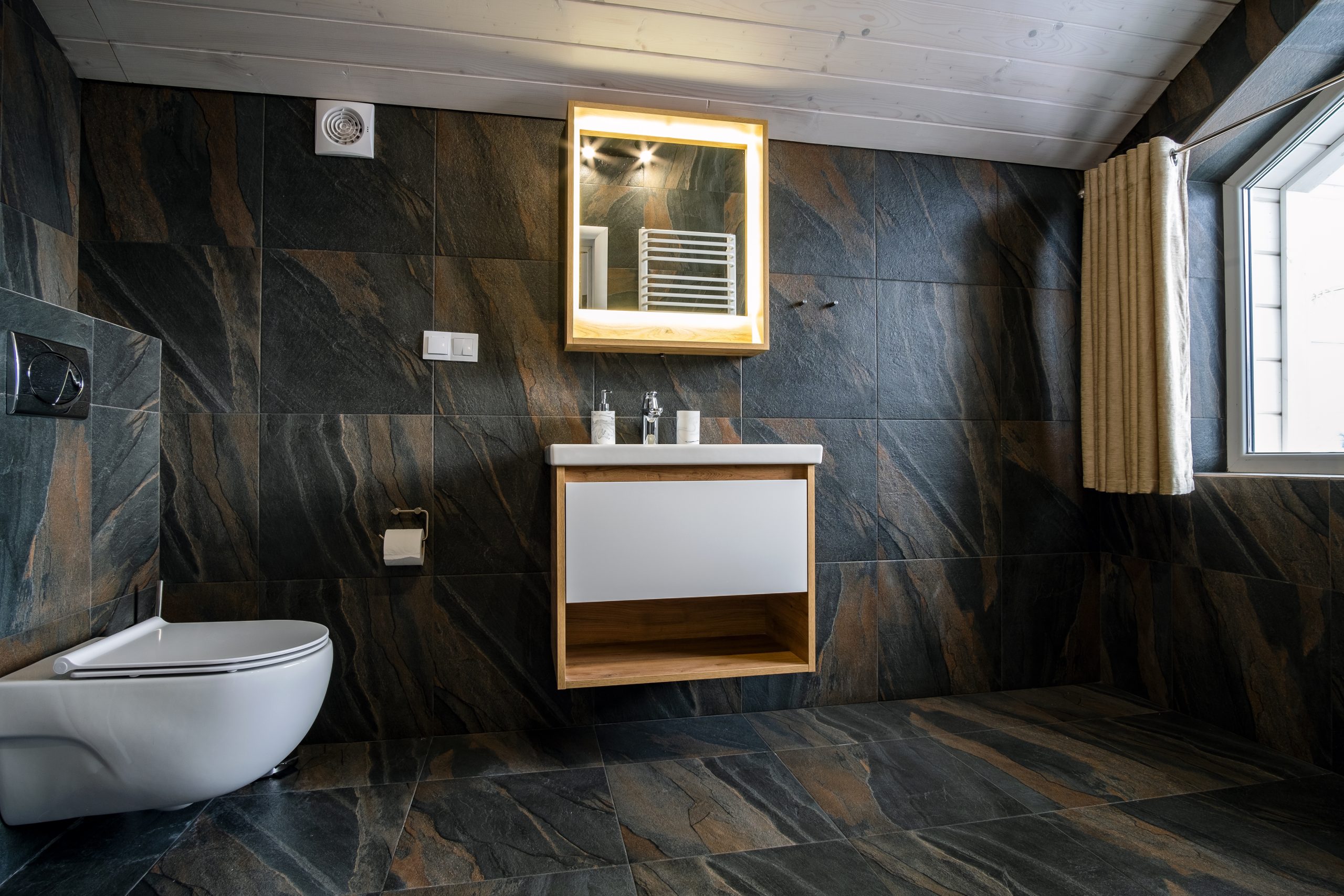 Interior of modern stylish bathroom with black tiled walls, curtain shower place and wooden furniture with wash basin and big illuminated mirror.