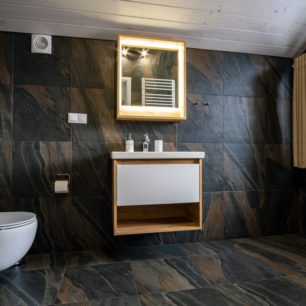 Interior of modern stylish bathroom with black tiled walls, curtain shower place and wooden furniture with wash basin and big illuminated mirror.